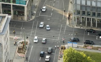 Ein Blick aus der Luft über eine große Kreuzung in einer Großstadt in der Mittagszeit, auf der viele Autos, Radfahrer und Fußgänger unterwegs sind.