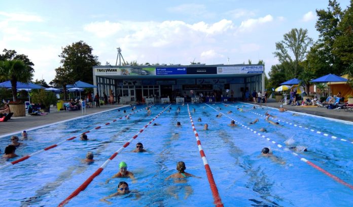 Das Sonnenbad in Karlsruhe. Die Badegäste schwimmen in den Becken und ziehen dort ihre Bahnen. Draußen ist es bewölkt – andere Gäste liegen und sitzen am Beckenrand am Wasser.