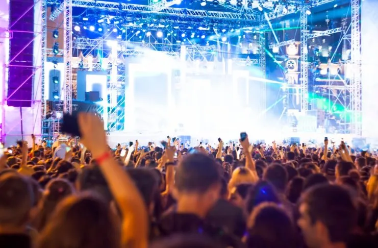 Zahlreiche Männer und Frauen stehen vor einer Open Air Bühne auf einem Festival und tanzen zu der Musik. Die Menschen auf der Veranstaltung freuen sich über die Show.