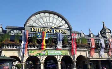Der Haupteingang zum Freizeitpark Europa-Park mit einer überdimensional großen kuppelförmigen Uhr, verschiedenen Flaggen Europas und mehreren Steinbögen. Direkt beim Eingang befinden sich die Kassen.