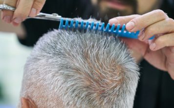 Ein Mann mit ergrautem Haar ist beim Friseur. Mit einem blauen Kamm und einer Haarschere schneidet die Friseurin die Haarspitzen des Herrn.