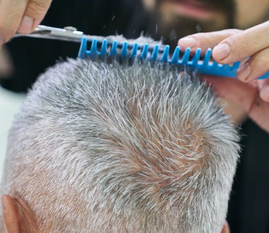 Ein Mann mit ergrautem Haar ist beim Friseur. Mit einem blauen Kamm und einer Haarschere schneidet die Friseurin die Haarspitzen des Herrn.