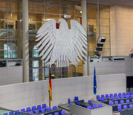 Den leeren Bundestag im Inneren mit blauen Sitzen und der Deutschlandflagge sowie der EU-Flagge im Vordergrund ziert auf einer großen Glasfront der Bundesadler.