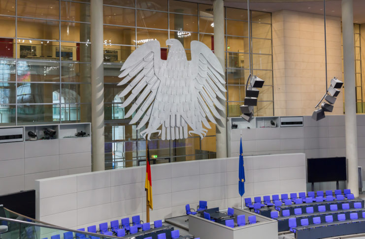 Den leeren Bundestag im Inneren mit blauen Sitzen und der Deutschlandflagge sowie der EU-Flagge im Vordergrund ziert auf einer großen Glasfront der Bundesadler.