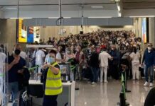 Es gibt einen großen Ansturm: Menschenmassen stehen in einem Flughafen an. Ordner in Sicherheitswesten versuchen die vielen Fluggäste zurück zu halten. Es droht ein Chaos.
