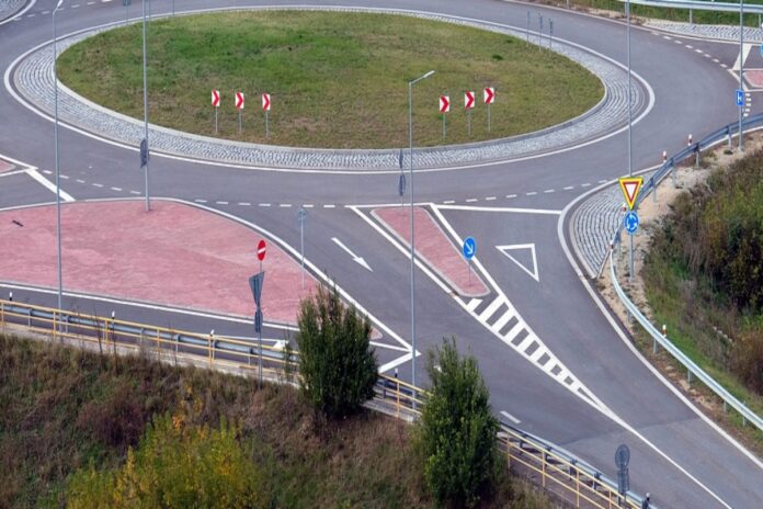 Ein großflächiger Kreisverkehr mit mehreren Spuren ist aus der Luftperspektive zu sehen. In der Mitte des nicht befahrenen Kreisels befindet sich eine Grasfläche und der Asphalt ist von Bäumen umgeben.
