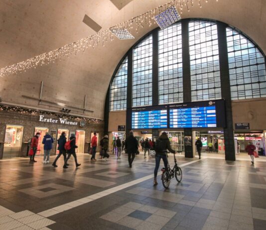 Eingangshalle des Hauptbahnhofs Karlsruhe. Fahrgäste durchqueren den Bahnhof und das Gebäude, um zu ihrem Zug oder Gleis zu gelangen. In der Mitte des Bahnhofs befindet sich eine große Anzeigetafel, auf der alle Abfahrtszeiten und Zugverbindungen auf einen Blick zu sehen sind.