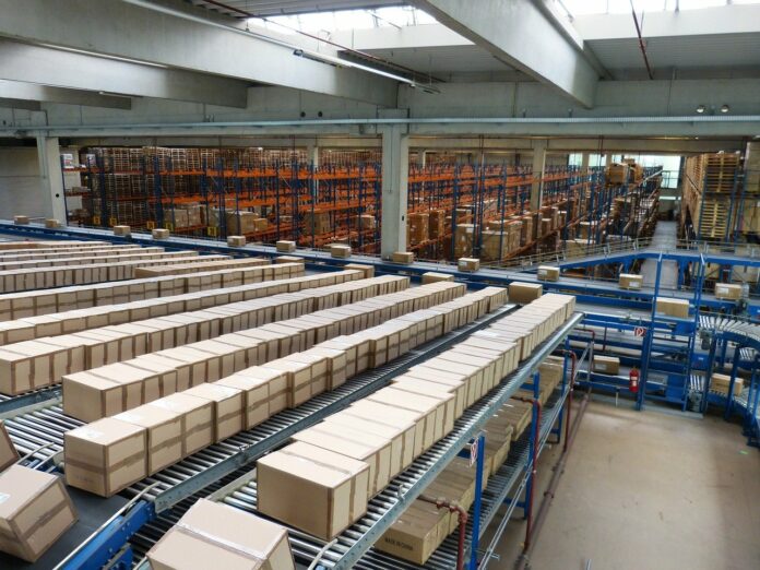 Auf dem Bild sieht man zahlreiche Pakete, die nebeneinander aufgereiht in Regalen eines Lagers einer Postzentrale liegen. Die Pakete warten darauf, von der DHL versendet zu werden.