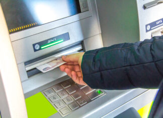 Eine Person steht vor dem Geldautomaten. Sie entnimmt das Bargeld aus dem Fach. Er hält die Hand über dem Zahlenfeld.