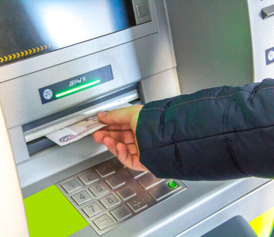 Eine Person steht vor dem Geldautomaten. Sie entnimmt das Bargeld aus dem Fach. Er hält die Hand über dem Zahlenfeld.