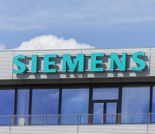 Das Siemens Geschäftsgebäude mit großem türkisem Schriftzug des Logos und des Namens. Darüber sieht man den strahlend blauer Himmel mit einigen Wolken.