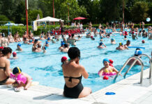 In einem Freibad tummeln sich viele Besucher und haben offensichtlich Spaß. Während sich die meisten im Becken befinden und schwimmen und spielen, sitzen andere am Beckenrand.