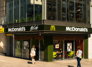 Ein modernes McDonald's-Fastfood-Restaurant in der Innenstadt an einer Ecke platziert. Es ist ebenfalls ein McCafé ausgeschildert. Die Filiale hat große Fenster.