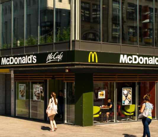 Ein modernes McDonald's-Fastfood-Restaurant in der Innenstadt an einer Ecke platziert. Es ist ebenfalls ein McCafé ausgeschildert. Die Filiale hat große Fenster.
