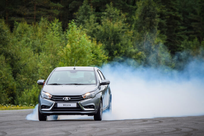 Ein silbernes Auto fährt über den Asphalt und qualmt sehr stark. Der blau-weiße Rauch umgibt den hinteren Teil des Lada Vesta. Die oberen Dreiviertel der Bildfläche im Hintergrund sind von grünen Bäumen eingenommen.