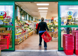 Eine Kundin hält einen Einkaufskorb in der Hand und geht mit schnellen Schritten einkaufen im Supermarkt. Am Eingang stehen Obst und Gemüse.