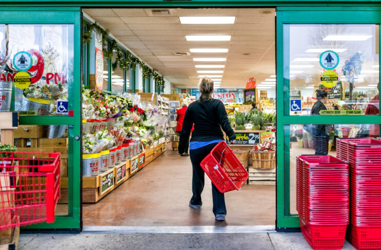 Eine Kundin hält einen Einkaufskorb in der Hand und geht mit schnellen Schritten einkaufen im Supermarkt. Am Eingang stehen Obst und Gemüse.