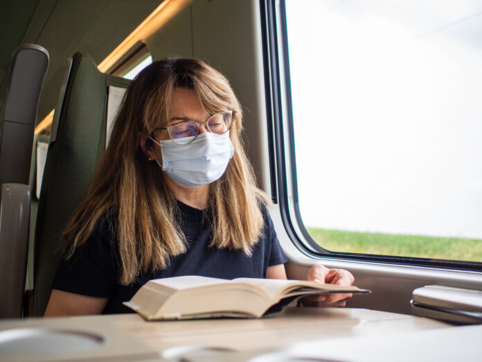 Frau mit Maske, fährt Zug, liest Buch