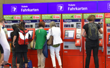 Ein roter Fahrkartenautomat am Bahnhof an dem mehrere Menschen Fahrkarten für die Bahn kaufen. Das 9-Euro-Ticket für Zugfahrt ist hier käuflich. An dem Automaten gibt es auch das Deutschlandticket für die Deutsche Bahn.