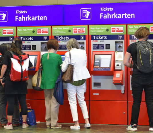 Ein roter Fahrkartenautomat am Bahnhof, an dem mehrere Menschen Fahrkarten für die Bahn kaufen. Das 9-Euro-Ticket für Zugfahrt ist hier käuflich. An dem Automaten gibt es auch das Deutschlandticket für die Deutsche Bahn.