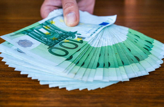 Die Hand einer Person, die mehrere 100-Euro-Scheine hält. Die vielen grünen Scheine, die auf einem braunen Tisch aufliegen, sind zu einem breiten, gleichmäßigen Fächer geformt.