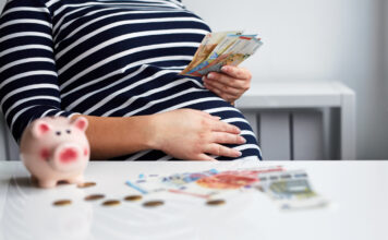 Eine schwangere Frau in einem gestreiften Oberteil, hält Geldscheine in der Hand, während auf dem Tisch weitere Scheine, Münzen und ein Sparschwein drapiert sind.