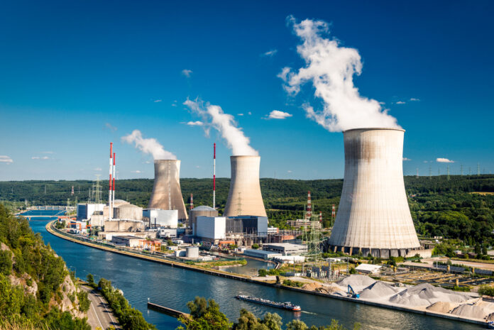 Zu sehen sind Atomkraftwerke, bestehend aus massiven grauen Kühltürmen, aus denen weißer Wasserdampf in den Himmel aufsteigt. Die Anlage liegt an einem Fluss, was auf die Nutzung von Wasser zur Kühlung hinweist. Die grüne Umgebung mit Wiesen und Bäumen steht im Kontrast zu dem industriellen Bauwerk.