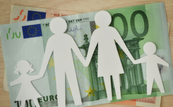 Eine ausgeschnittene Familie aus weißem Papier liegt auf einem 50-Euro- und einem 100-Euro-Schein. Die Familie besteht aus Mutter, Vater, Sohn und Tochter.