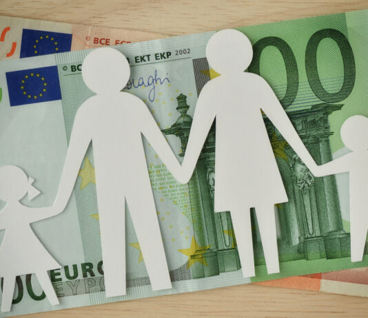 Eine ausgeschnittene Familie aus weißem Papier liegt auf einem 50-Euro- und einem 100-Euro-Schein. Die Familie besteht aus Mutter, Vater, Sohn und Tochter.