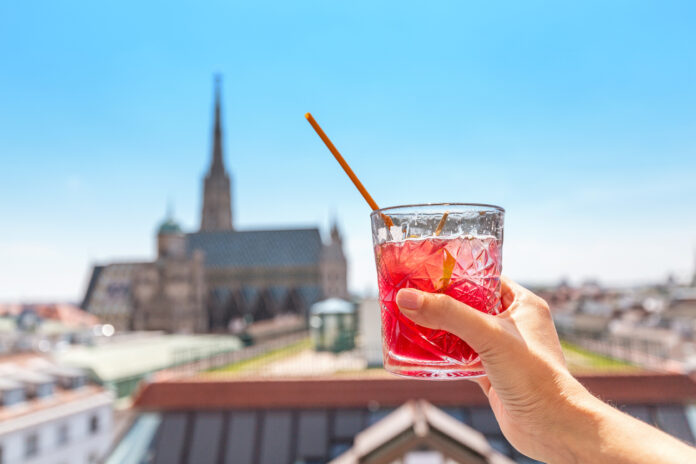 Ein Gast hält ein Glas mit einem Getränk in der Hand, das ein Kellner in einer Bar an den Tisch gebracht hat. Die Terrasse bietet einen Blick über die ganze Stadt.