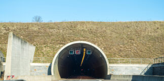 Einfahrt in einen dunklen Tunnel oder Autobahntunnel mit Beschilderung, Verkehrszeichen, Beleuchtung und Ampeln. Die Straße, die durch den Tunnel führt, ist zweispurig.