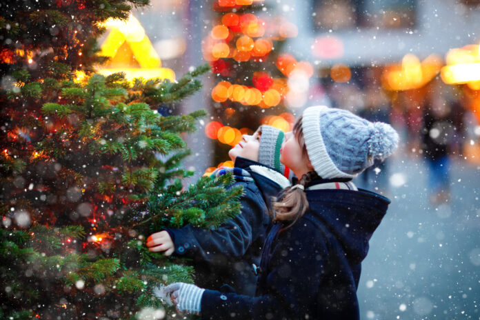 Zwei Kinder stehen bei Schnee vor einem Weihnachtsbaum.