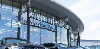Eine Reihe polierter, schwarzer Neuwagen parken im Sonnenlicht vor der Glasfassade einer modernen Mercedes Benz Filiale. Die Luxusfahrzeuge präsentieren sich im Vordergrund vor dem Mercedes-Benz Logo.