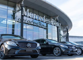 Eine Reihe polierter, schwarzer Neuwagen parken im Sonnenlicht vor der Glasfassade einer modernen Mercedes Benz Filiale. Die Luxusfahrzeuge präsentieren sich im Vordergrund vor dem Mercedes-Benz Logo.