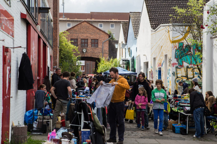 In einer Fußgängerzone einer Altstadt tummeln sich zahlreiche Menschen und Passanten. Sie gehen shoppen und einige schauen sich einen Secondhand-Laden oder Vintage Store und seine Auswahl genauer an.