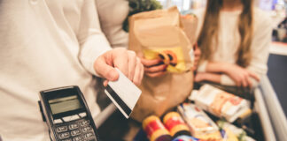 Eine Frau zahlt an der Supermarktkasse ihren üppigen Einkauf mit Karte. Während die Lebensmittel am Band liegen und sich eine Schlange hinter ihr bildet, hält sie die EC-Karte an das Kartenlesegerät vor.
