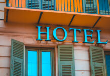 Nahaufnahme eines Schriftzugs mit dem Wort "Hotel". Es handelt sich dabei offenbar um einen Gasthof. Über dem Schriftzug sieht man einen Balkon mit geschlossenen Fensterläden. Es sieht sauber und ordentlich aus. Die Farben des Gebäudes in Orange und Türkis laden zum Übernachten und Verweilen ein.