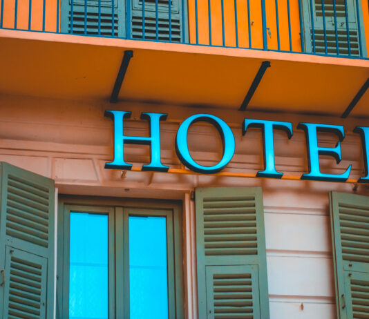 Nahaufnahme eines Schriftzugs mit dem Wort "Hotel". Es handelt sich dabei offenbar um einen Gasthof. Über dem Schriftzug sieht man einen Balkon mit geschlossenen Fensterläden. Es sieht sauber und ordentlich aus. Die Farben des Gebäudes in Orange und Türkis laden zum Übernachten und Verweilen ein.