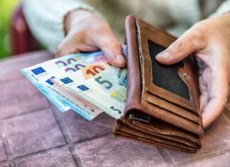 Die Hände eines alten Menschen halten einen Geldbeutel aus Leder, in dem mehrere unterschiedliche Geldscheine stecken. Die Hände ruhen auf einem vergilbten Gartentisch.