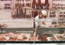 Eine gut bestückte Fleischtheke im Supermarkt. Ein Mitarbeiter nimmt die Waren aus der Kühltheke und im Hintergrund hängen allerhand Wurstwaren und Fischereiprodukte bereit.