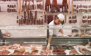 Eine gut bestückte Fleischtheke im Supermarkt. Ein Mitarbeiter nimmt die Waren aus der Kühltheke und im Hintergrund hängen allerhand Wurstwaren und Fischereiprodukte bereit.