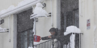 Ein Mann ist auf einem Balkon und versucht, eine Fensterscheibe von Eis zu befreien. Es ist tiefster Winter und der Balkon ist zugeschneit. Der Mann trägt eine dicke Winterjacke und noch mehr Schneeflocken fallen vom Himmel.