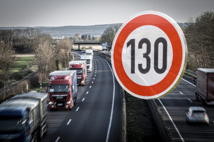Eine viel befahrene Autobahn – in Deutschland oder einem Nachbarland – schlängelt sich durch die Landschaft. Auf einer Spur fahren sehr viele Lkw. Am rechten Bildrand zeigt ein Tempo-130-Schild, dass hier ein Tempolimit gilt.