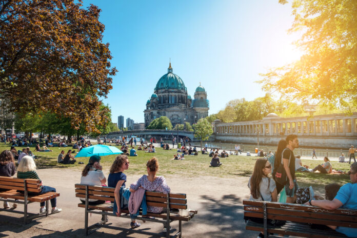 Menschen sitzen im Park. Es ist ein schöner, warmer und sonniger Tag. Die Menschen tragen T-Shirts und sitzen auf Parkbänken oder im Gras. Im Hintergrund sieht man einen Fluss. Die Blätter an den Bäumen sind leicht verfärbt.