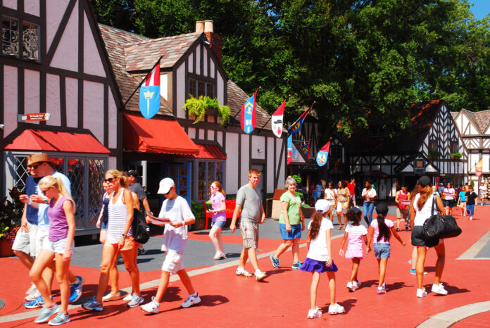 Zahlreiche Gäste laufen im Sommer und in entsprechend sommerlicher Kleidung in einem Freizeitpark umher. Im Hintergrund stehen Häuser im Fachwerkhaus-Stil. Es wehen Fahnen im Wind und orangefarbene Markisen sind herabgelassen.