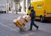 Ein Postzusteller von DHL läuft mit einem Wagen voller Pakete der Deutschen Post über eine Straße. Sein Auto steht am Straßenrand.