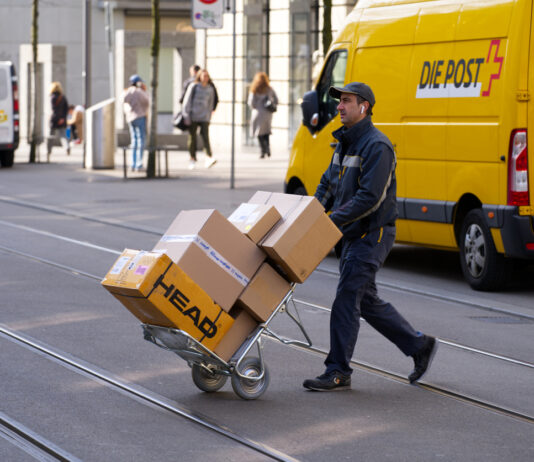 Ein Postzusteller von DHL läuft mit einem Wagen voller Pakete der Deutschen Post über eine Straße. Sein Auto steht am Straßenrand.