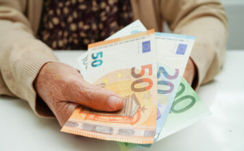 Eine Seniorin sitzt am Tisch und hält Geldscheine in der Hand und trägt dabei eine Strickjacke und ein geblümtes Oberteil.