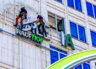 Das grüne Logo einer Galeria Kaufhof Filiale wird abgenommen. Zwei Bauarbeiter sind gesichert und schweben an der Wand des Gebäudes.