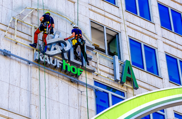 Das grüne Logo einer Galeria Kaufhof Filiale wird abgenommen. Zwei Bauarbeiter sind gesichert und schweben an der Wand des Gebäudes.